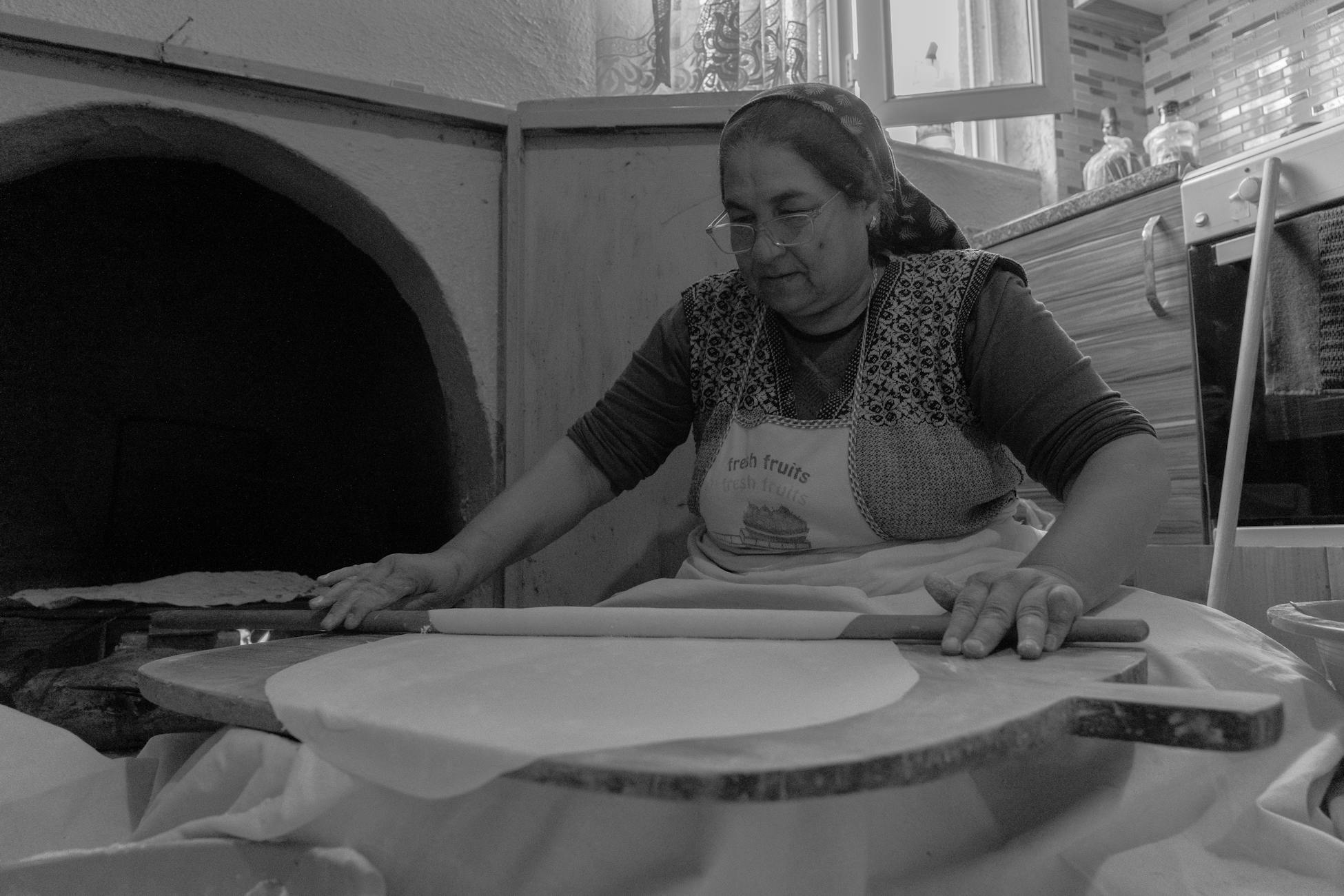 A Turkish woman making flatbread indoors with a rolling pin near a traditional oven.