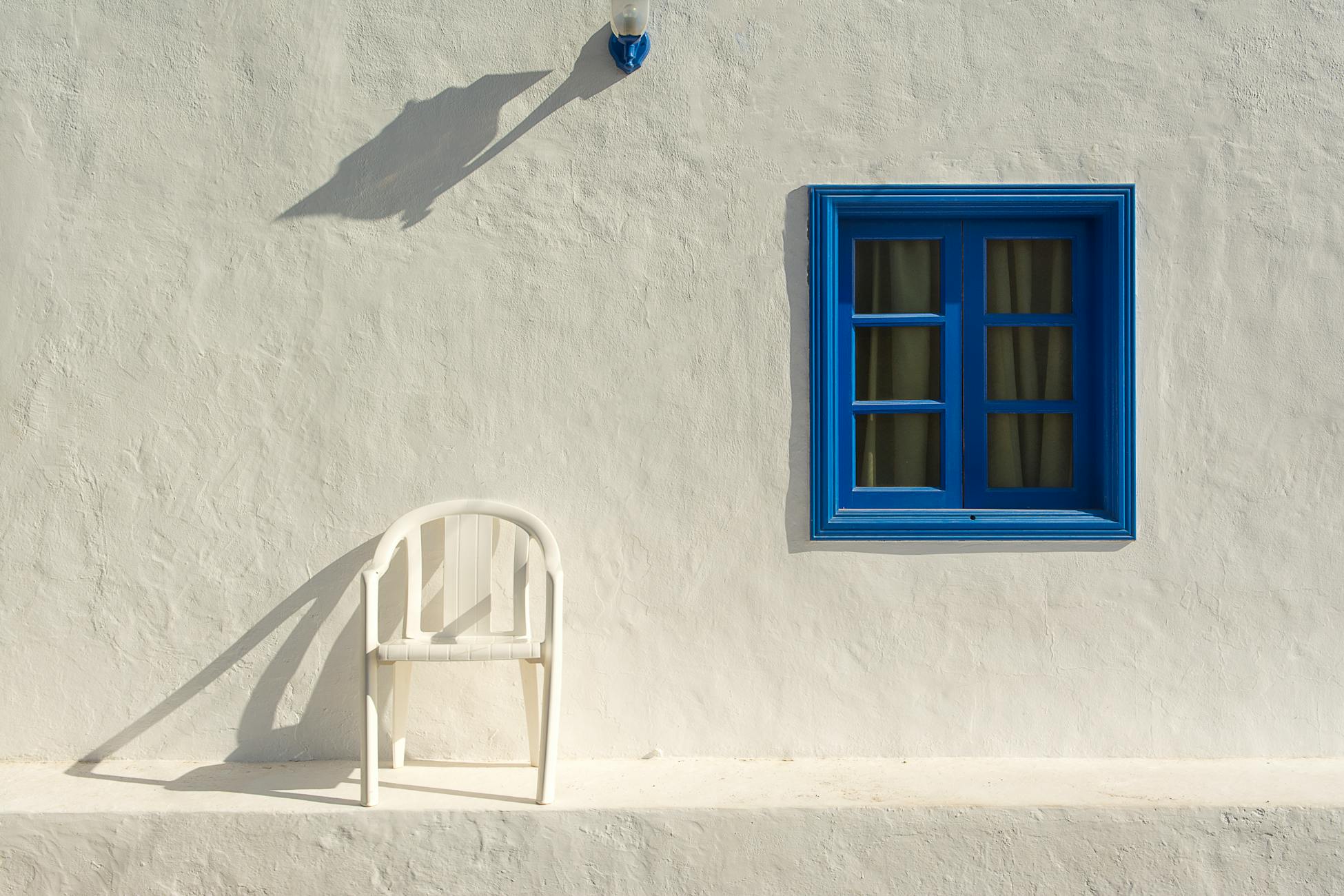 A stark white wall with a blue window frame and a white plastic chair casting shadows.