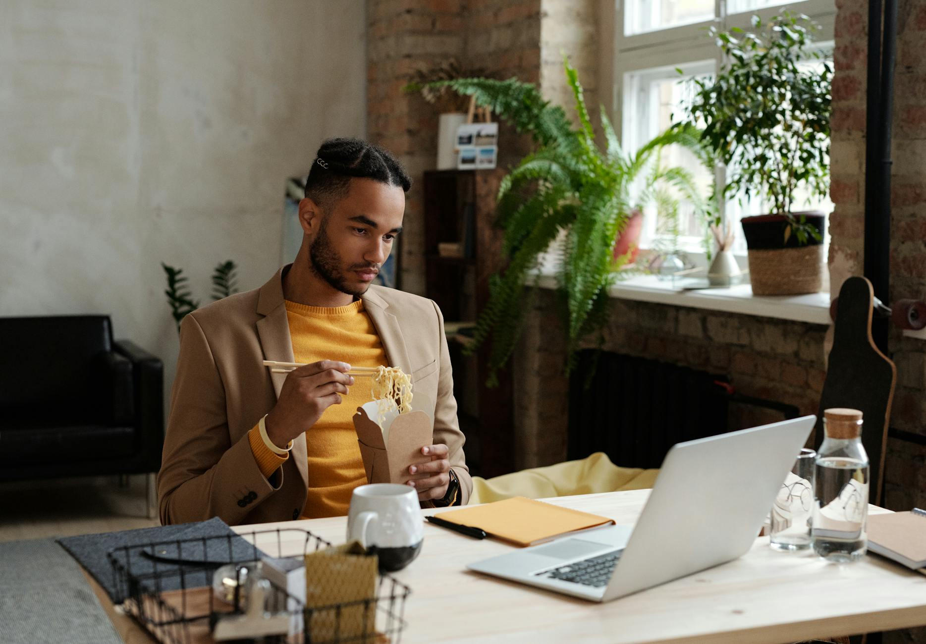 A young man enjoying takeout noodles while working at a modern office desk.