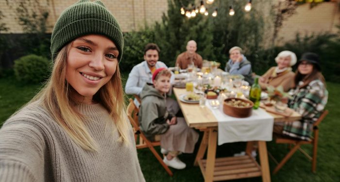 A joyful family dinner outdoors, captured with a smiling woman in the foreground taking a selfie.