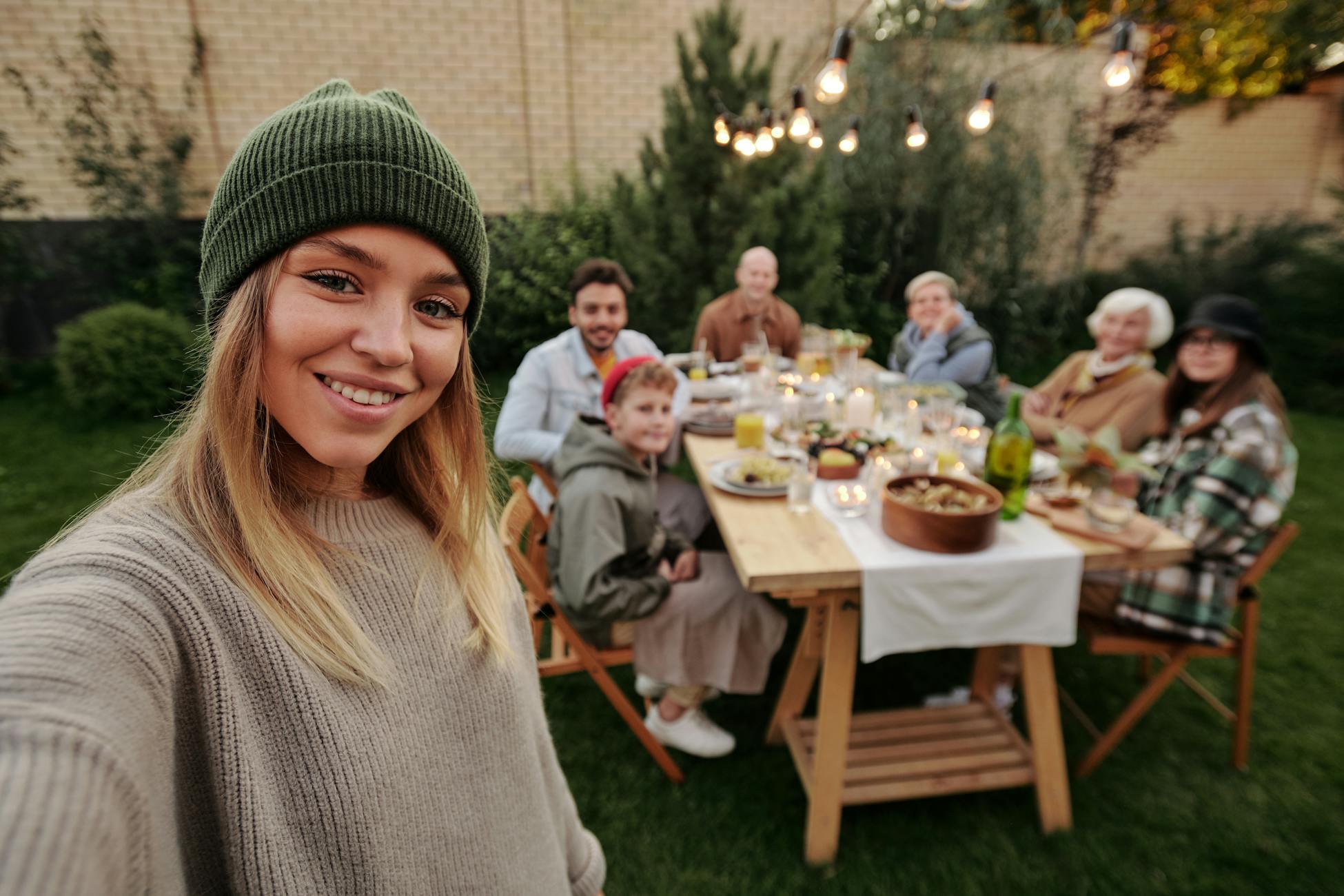 A joyful family dinner outdoors, captured with a smiling woman in the foreground taking a selfie.
