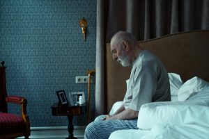 Elderly man sitting on bed, reflecting, in a cozy classic bedroom setting.