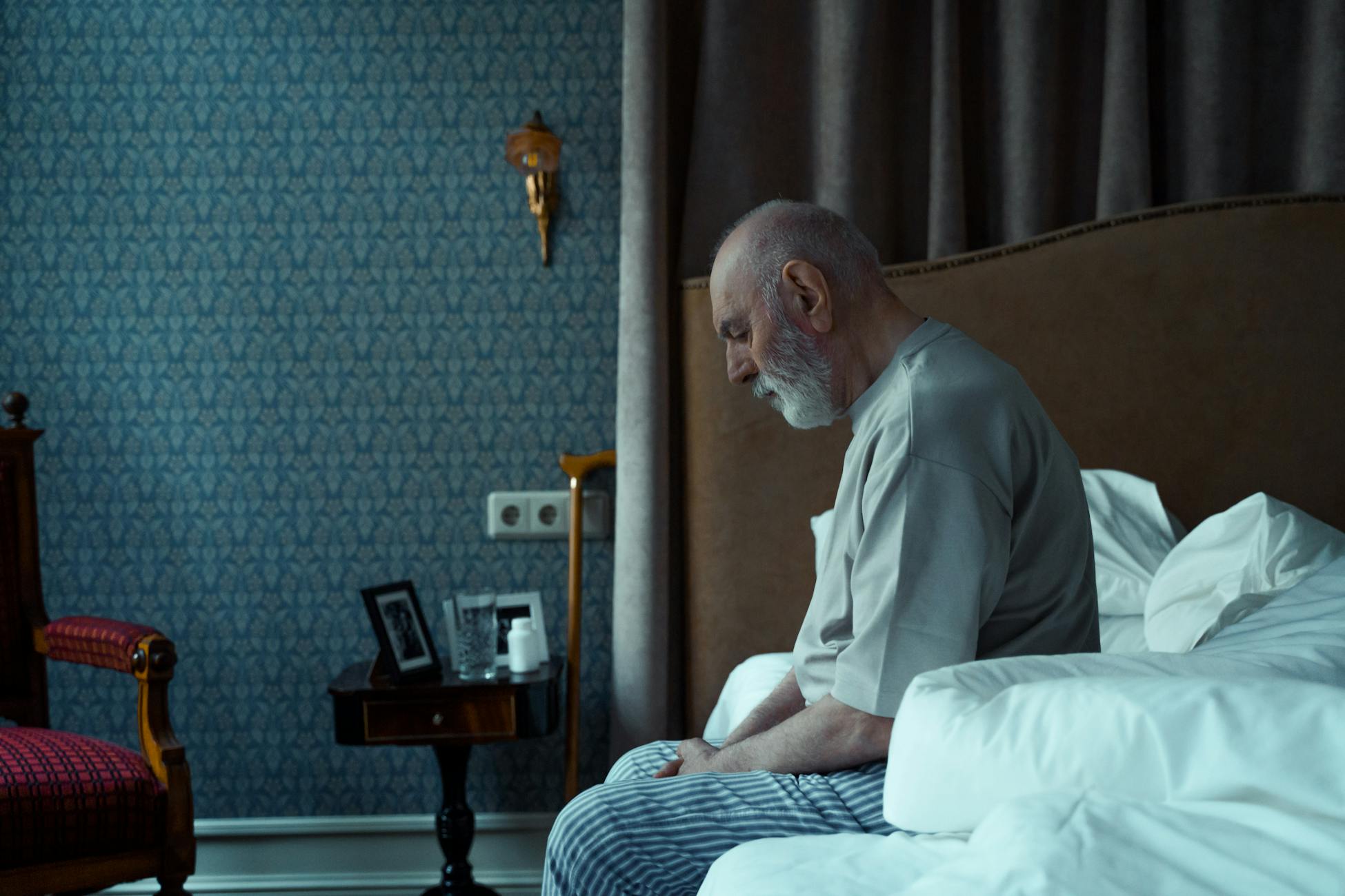 Elderly man sitting on bed, reflecting, in a cozy classic bedroom setting.