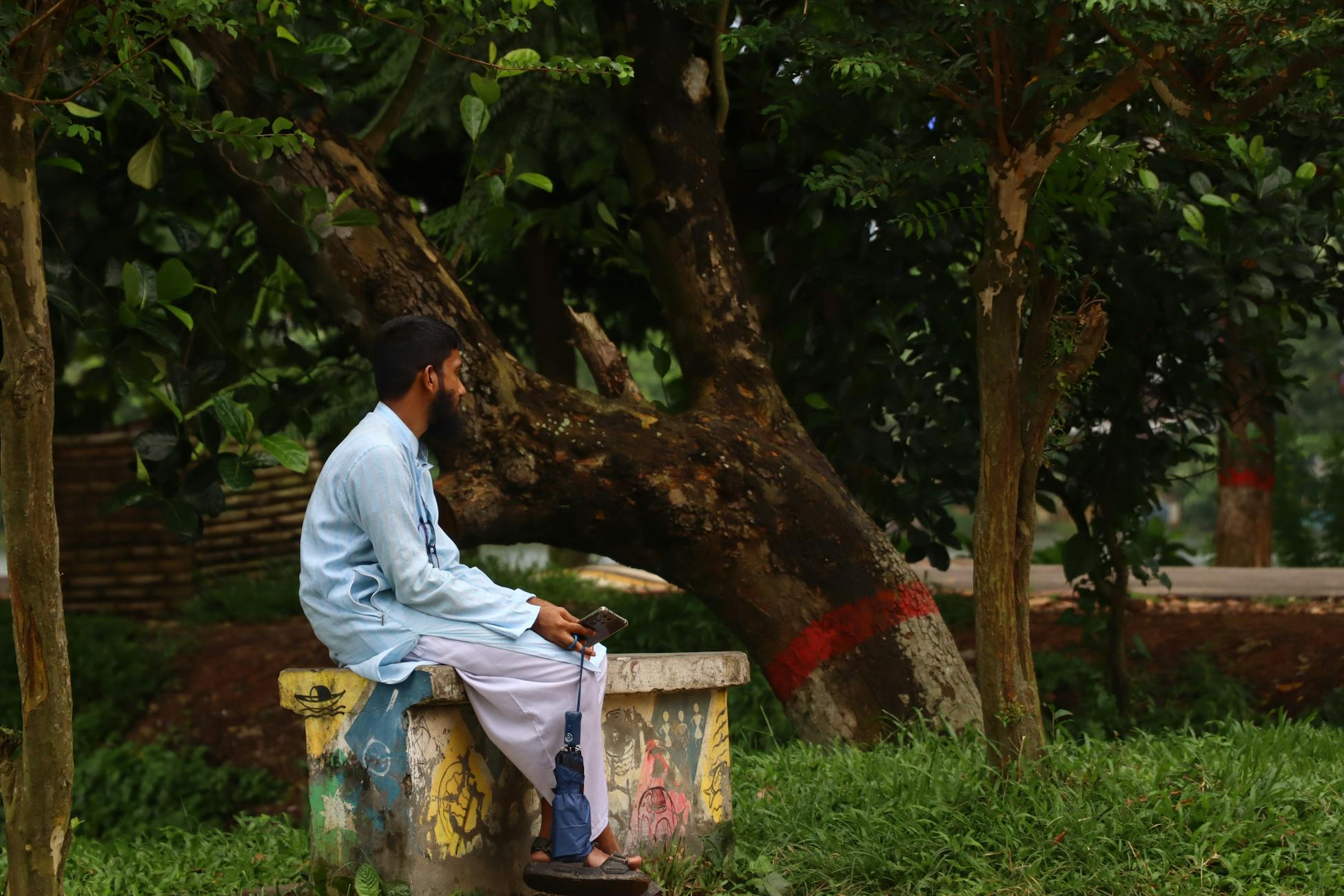 Man in traditional attire sitting on a bench in a tranquil park with lush greenery.