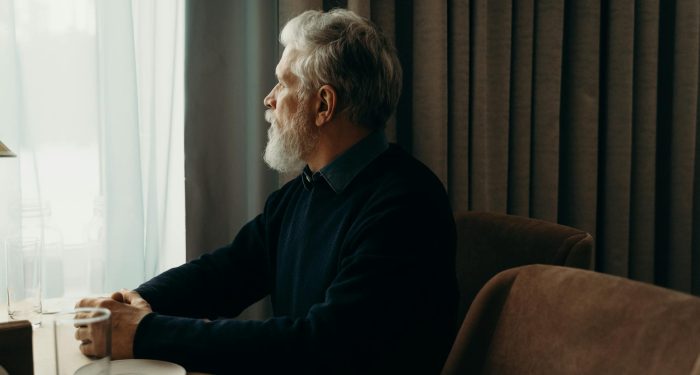 An elderly man with a beard looking outside while sitting by a table indoors.