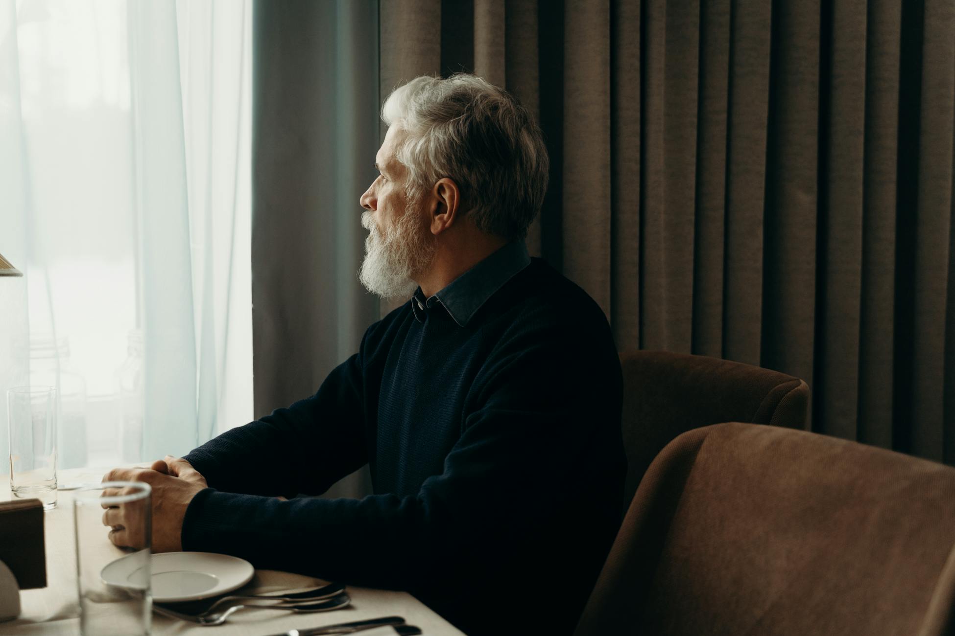 An elderly man with a beard looking outside while sitting by a table indoors.