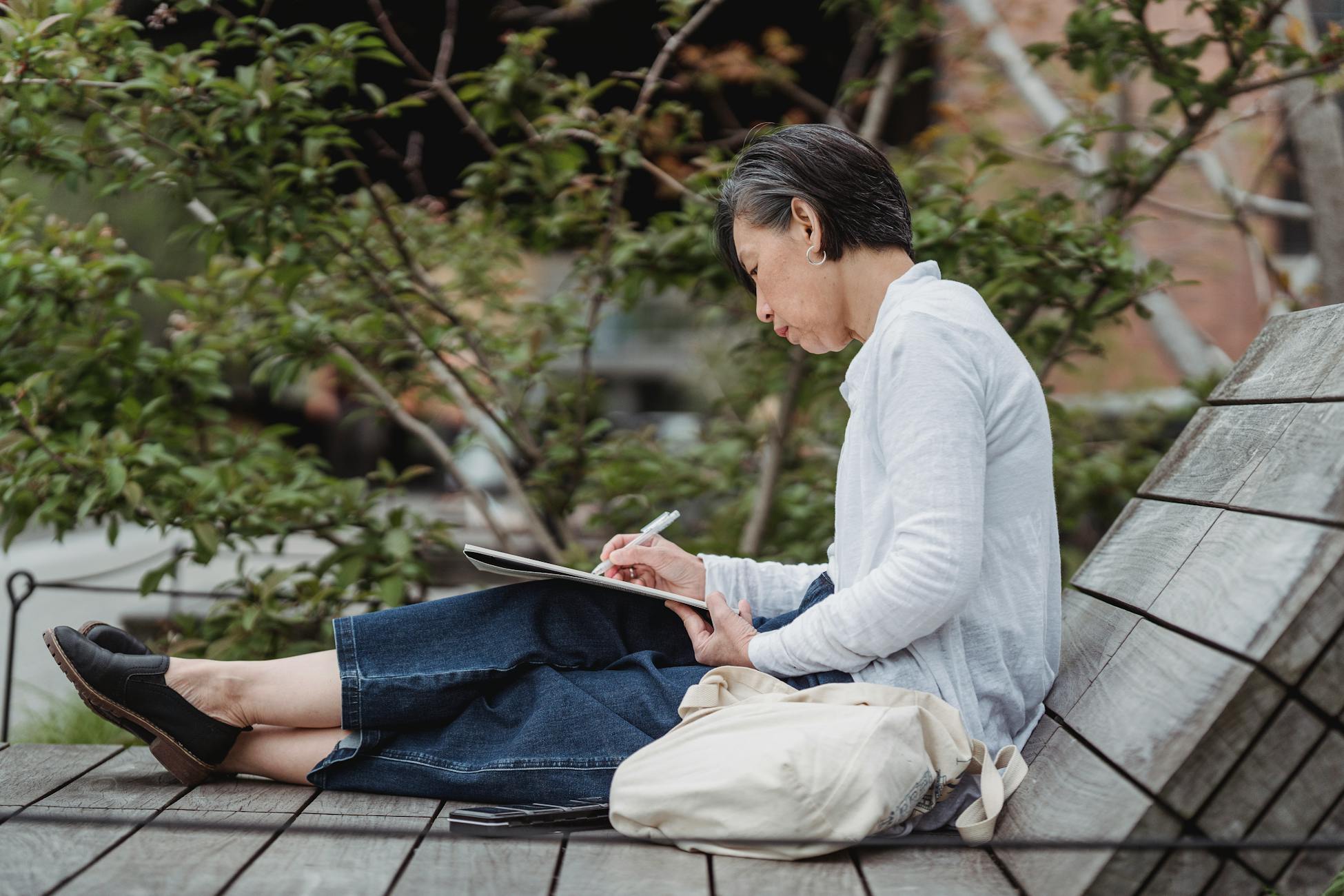A woman sits on a bench in an urban park, sketching in a notebook. Side view art scene.