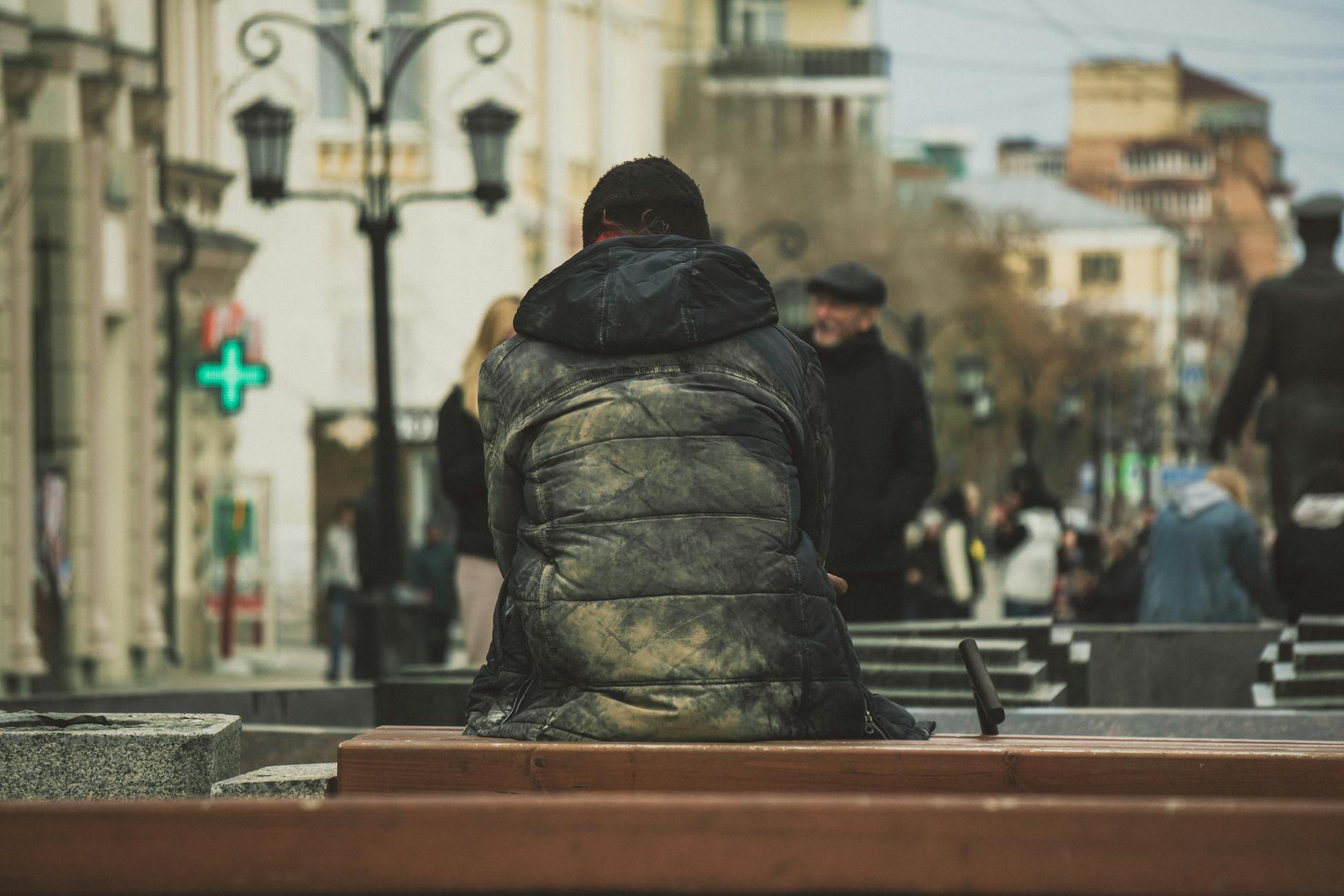 A solitary man in a puffer jacket sits on a bench in a bustling city street, observing passersby.