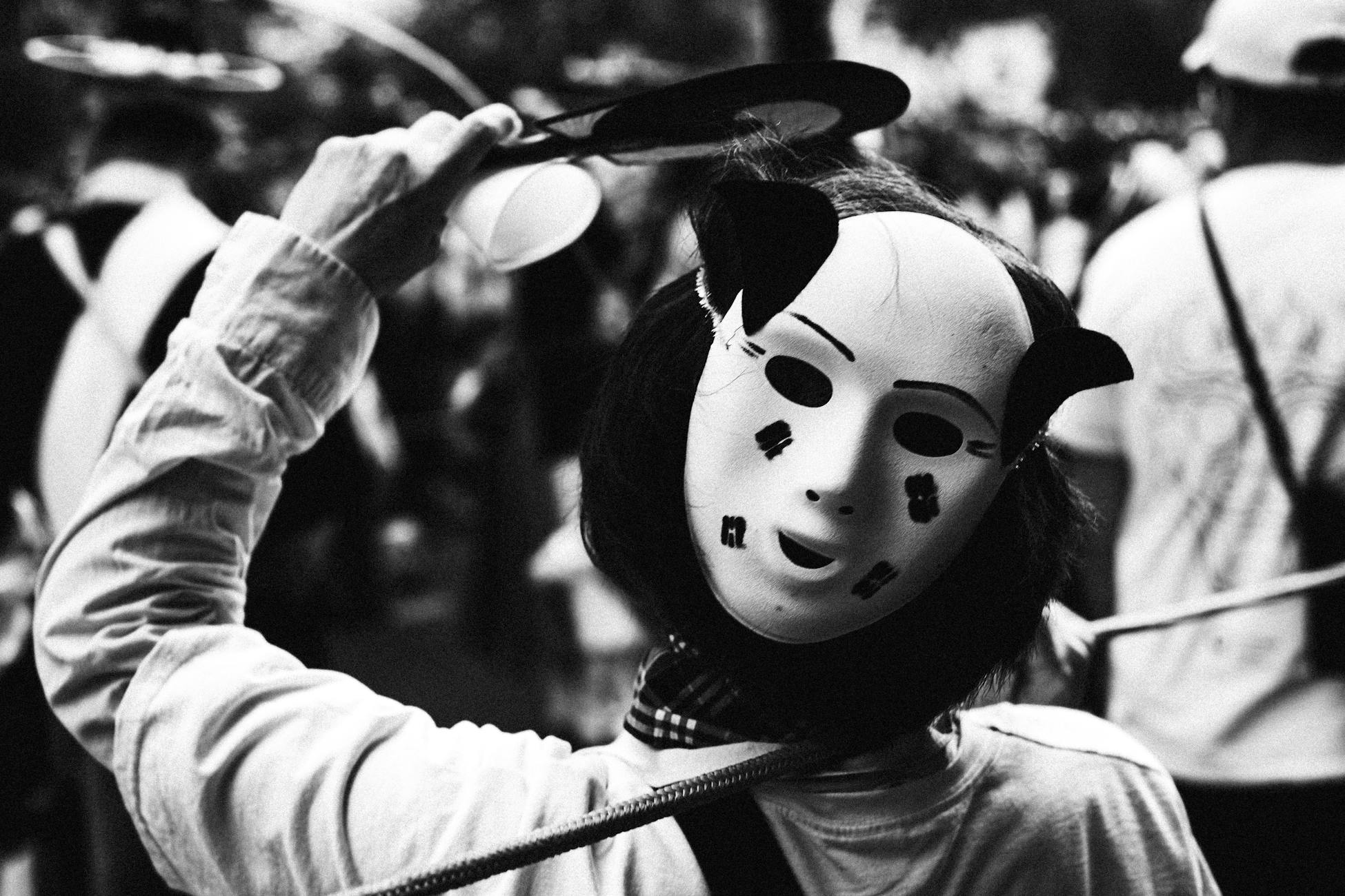 Black and white image of a masked figure surrounded by a bustling crowd at a Berlin festival.
