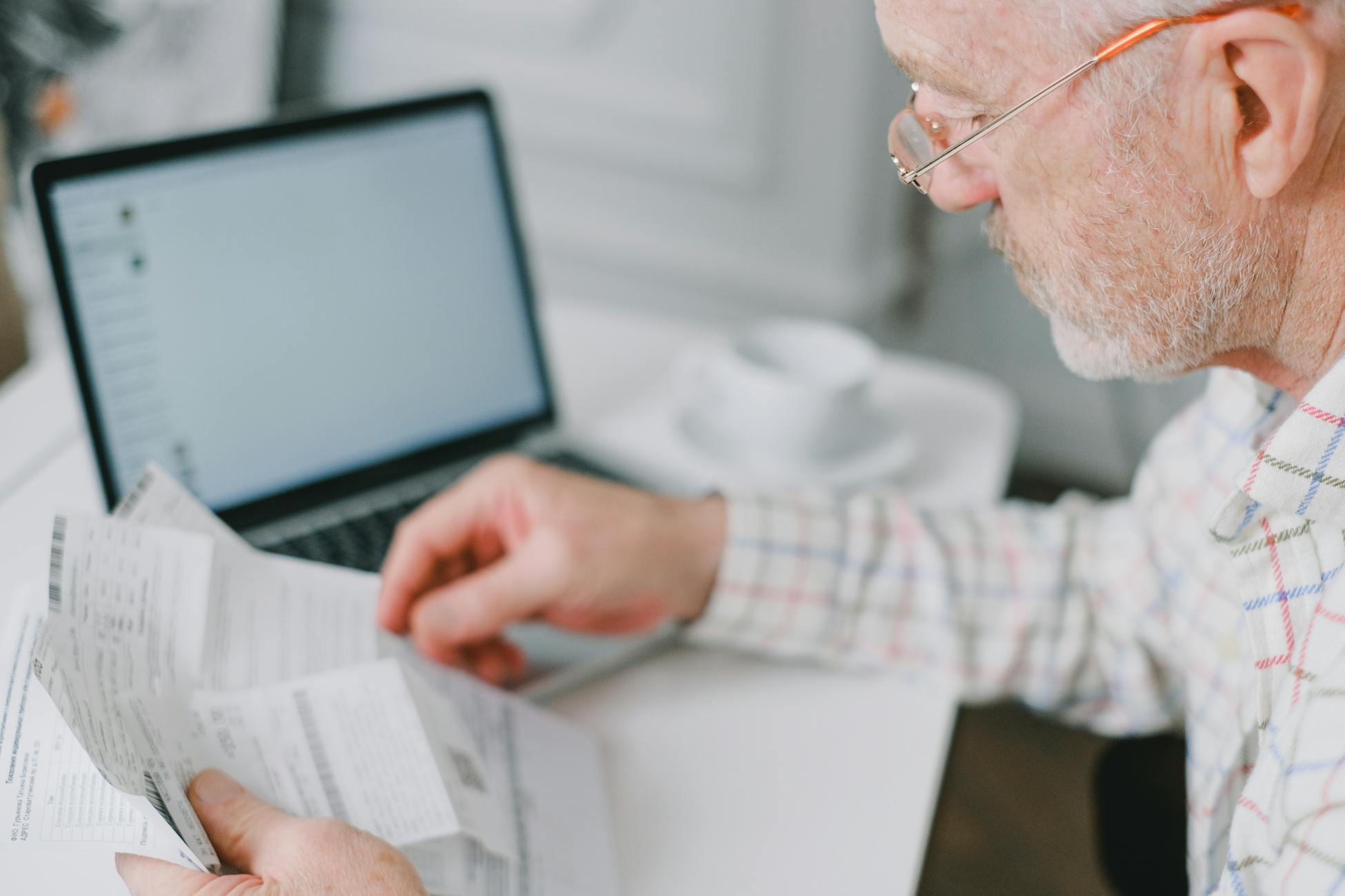 Elderly man wearing eyeglasses reading documents next to a laptop at home.