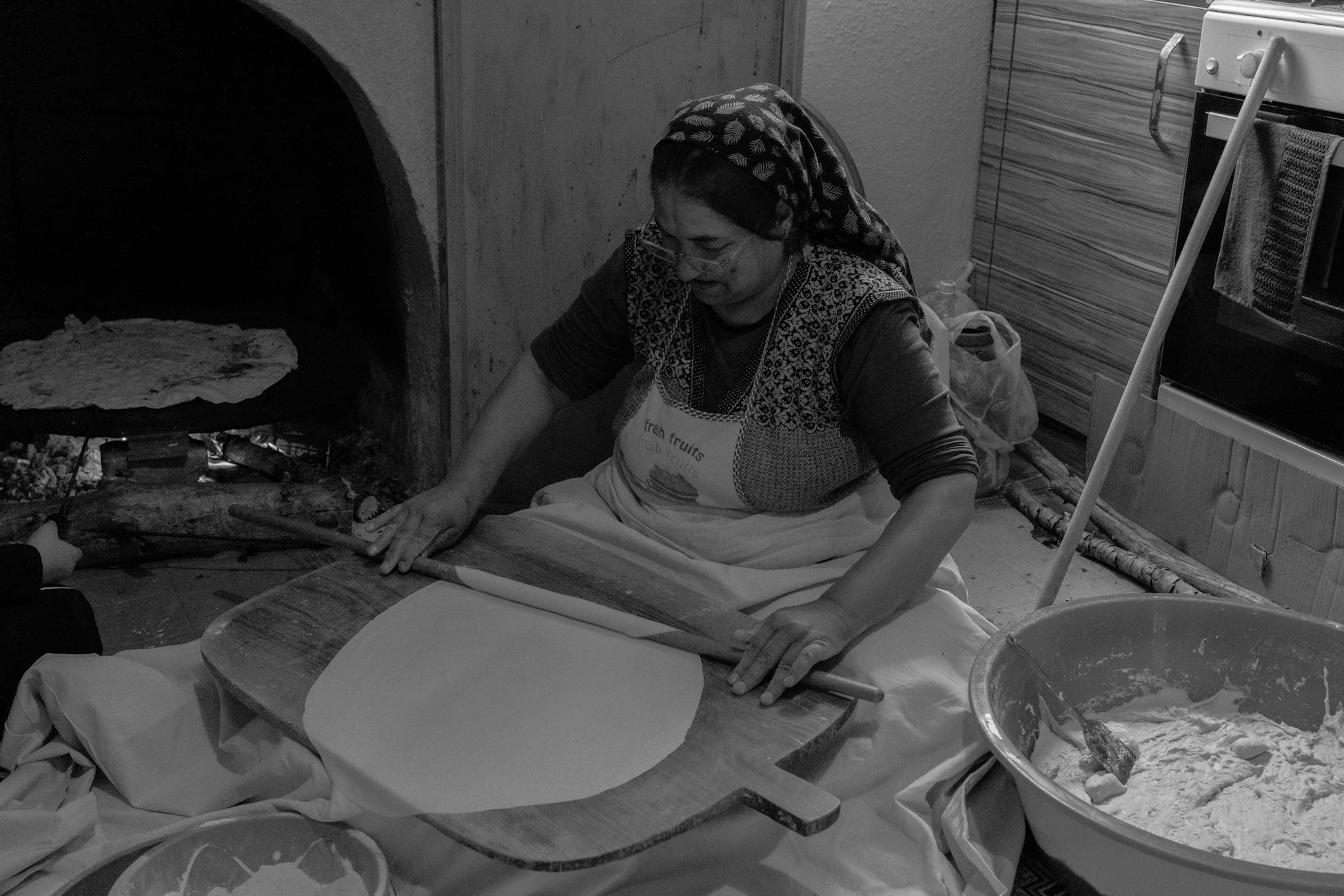An older woman making traditional Turkish flatbread indoors with a wood-fired oven.