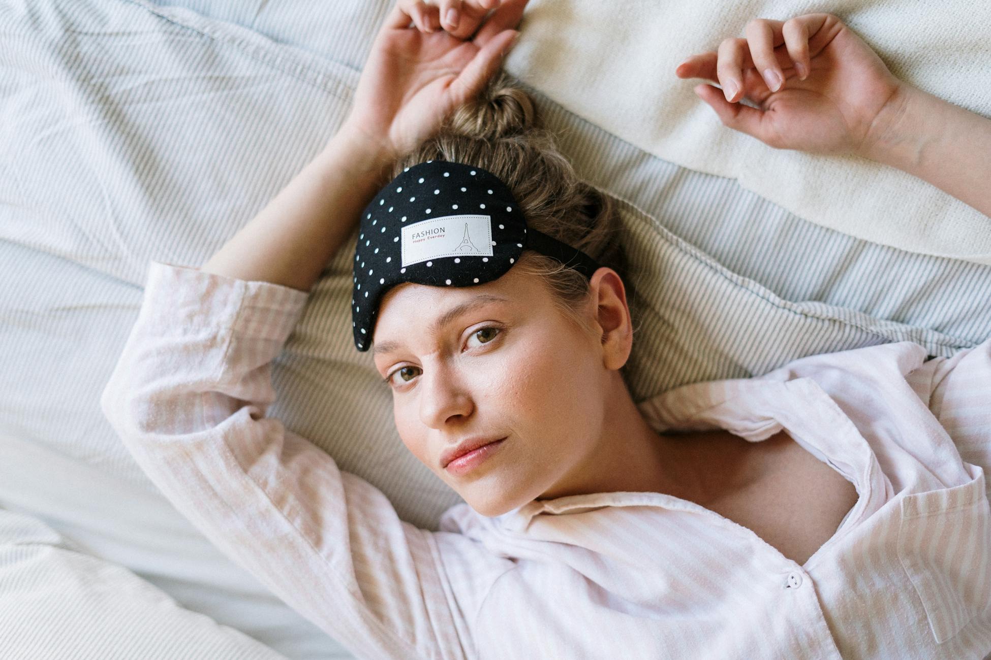 A young woman in pajamas and eye mask wakes up comfortably in bed, enjoying a relaxing morning.