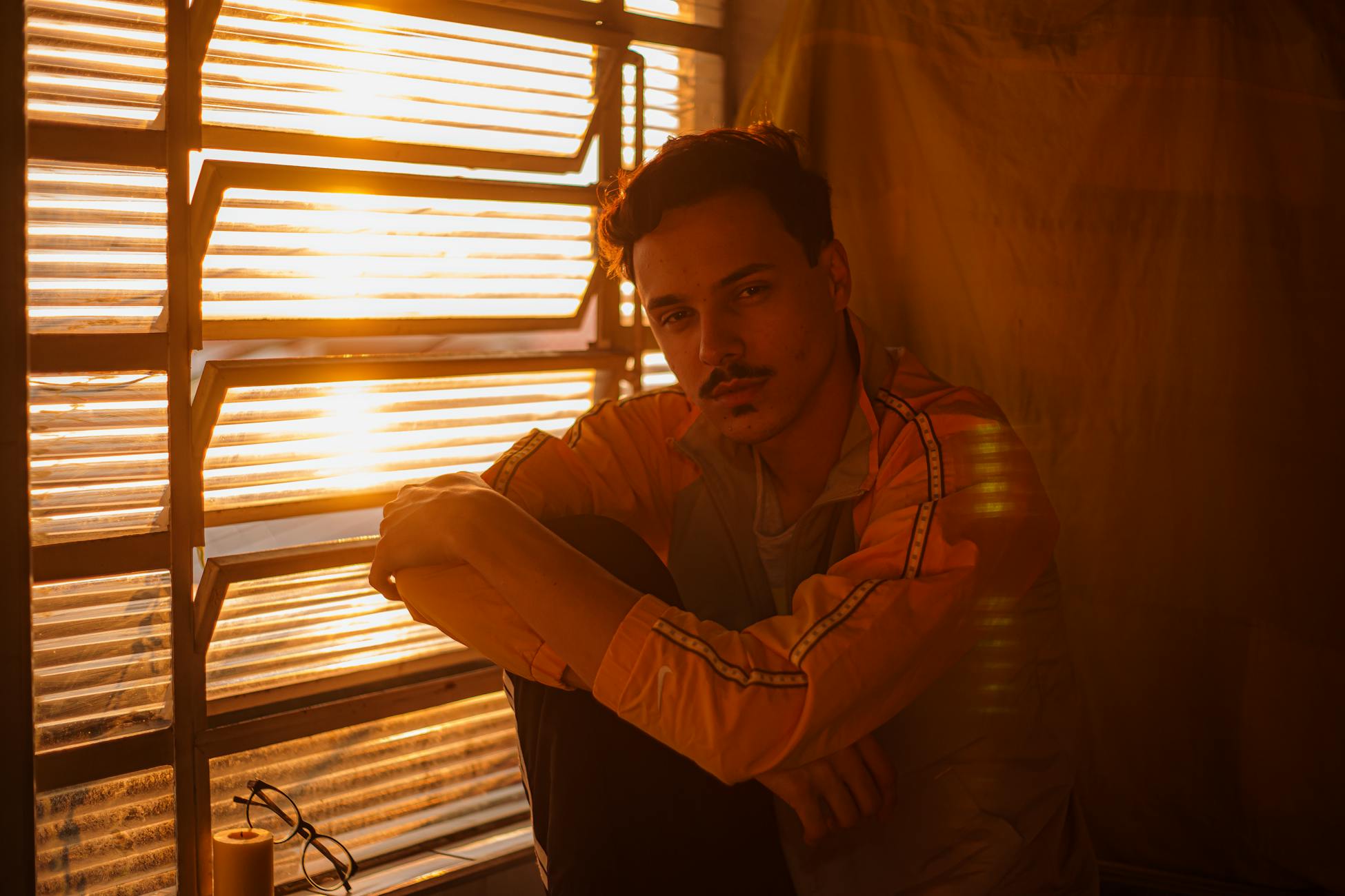 A young man sits thoughtfully by a window with warm sunlight streaming in, creating a serene mood.