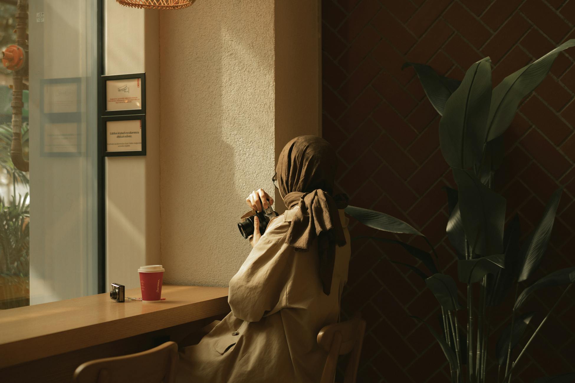 A woman in a hijab enjoys coffee while holding a camera in a cozy café setting.