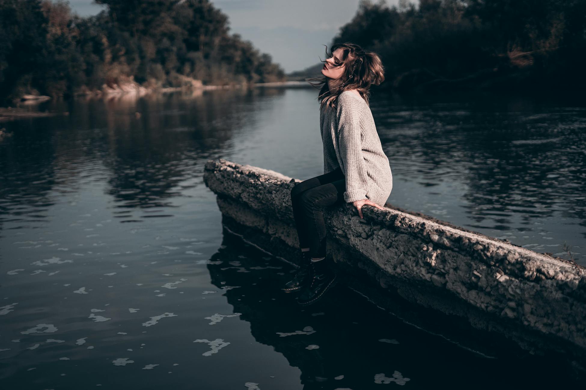 A woman enjoys a peaceful moment sitting on a riverside barrier during sunset.
