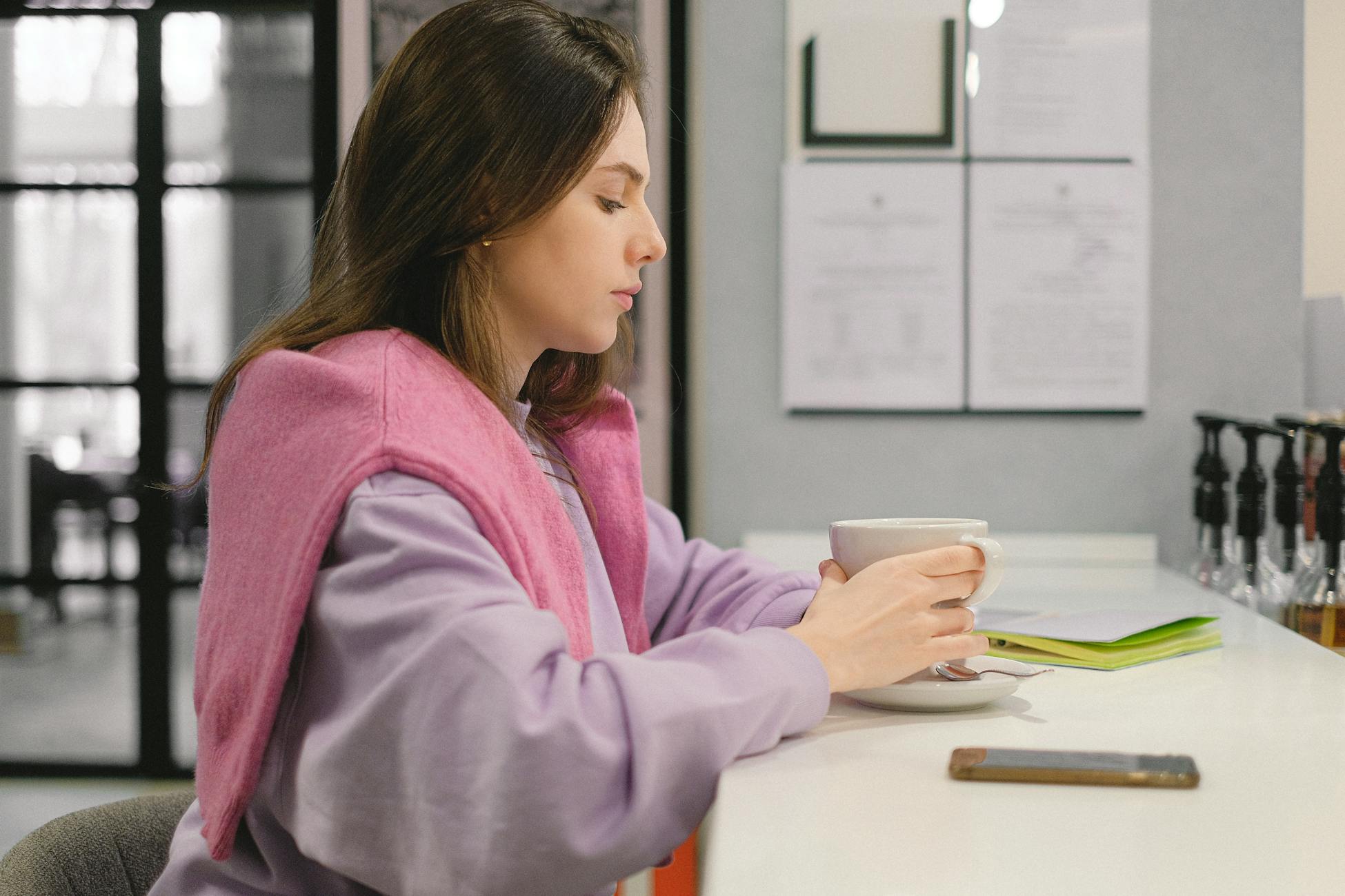 Woman enjoying a warm drink indoors, reflecting while holding a cup at a cafe counter.