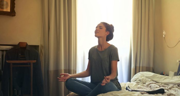 Adult woman practicing meditation on her bed surrounded by a calm bedroom atmosphere.