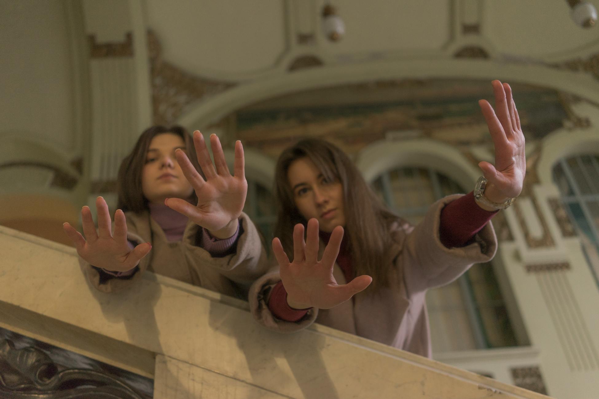 Two young women reach out their hands indoors in a historical setting.
