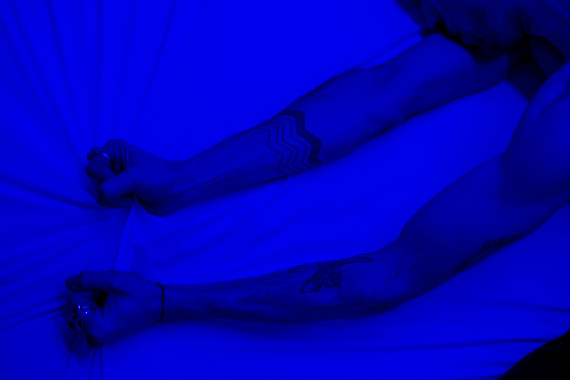 Close-up of tattooed arms grasping a white bedsheet under blue lighting, depicting anxiety and tension.