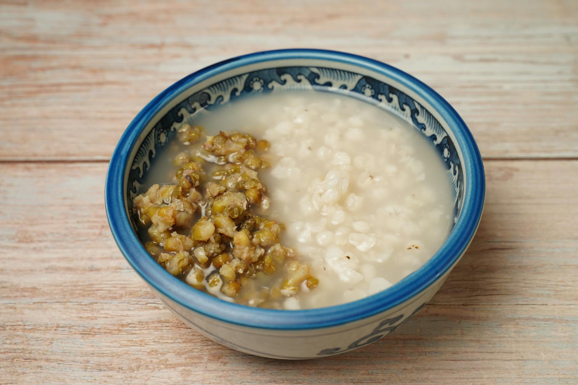 A close-up of mung bean and rice porridge served in an ornate blue and white ceramic bowl on a wooden surface.