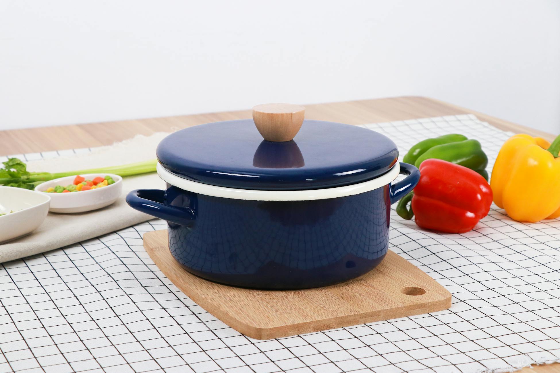 A blue pot on a wooden board surrounded by bell peppers and vegetables on a checkered tablecloth.