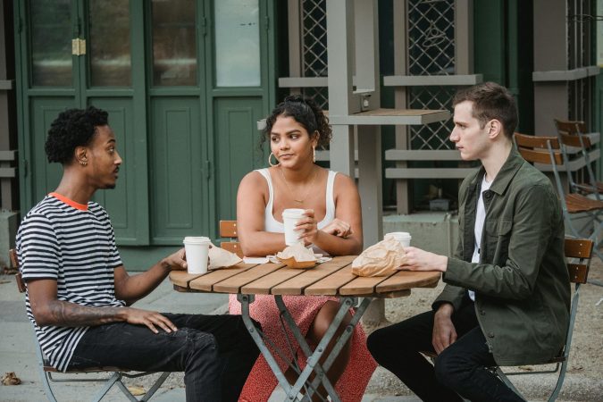 Young multiethnic friends in casual outfits drinking takeaway coffee and eating pastries while sitting at wooden table in street cafe