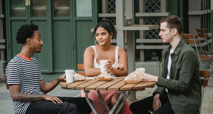 Young multiethnic friends in casual outfits drinking takeaway coffee and eating pastries while sitting at wooden table in street cafe