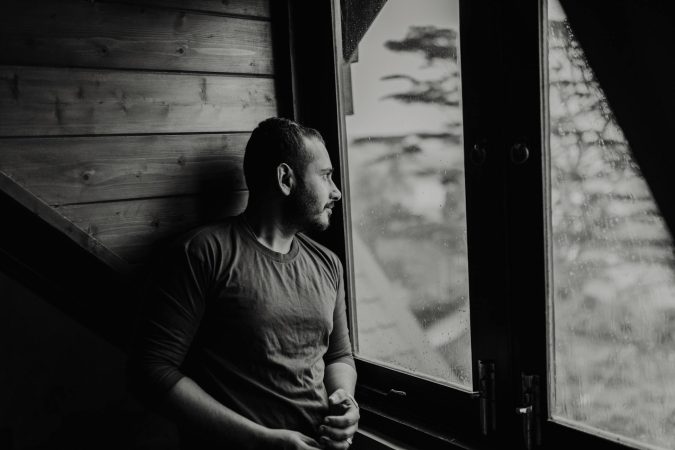 A bearded man thoughtfully gazes outside a window on a rainy day.