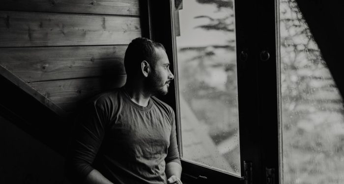 A bearded man thoughtfully gazes outside a window on a rainy day.