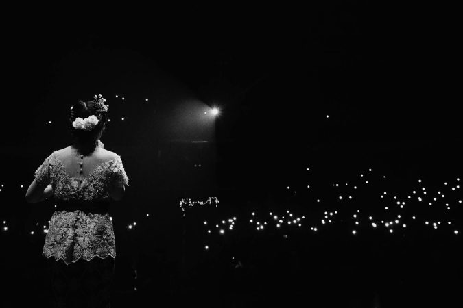A woman performer stands on stage illuminated by glowing audience lights in a dramatic black and white scene.