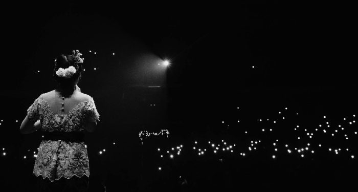 A woman performer stands on stage illuminated by glowing audience lights in a dramatic black and white scene.