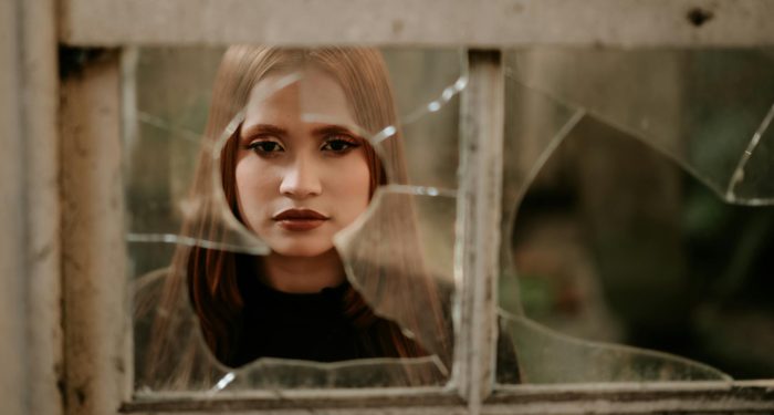 A young woman gazes solemnly through a shattered window, invoking emotions of solitude and introspection.