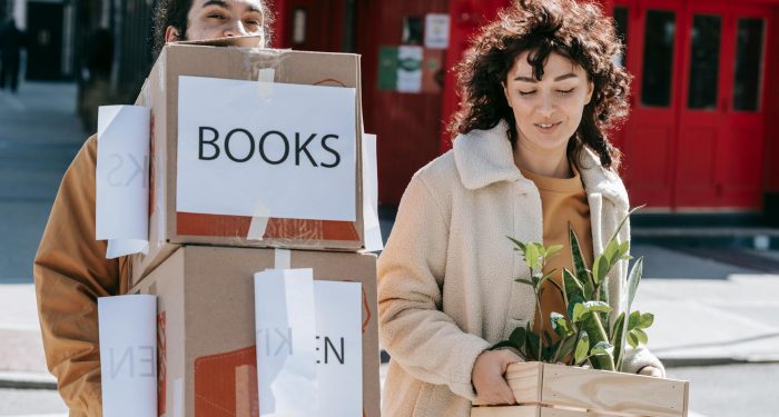 A joyful couple carrying boxes and plants while moving into a new home in an urban setting.
