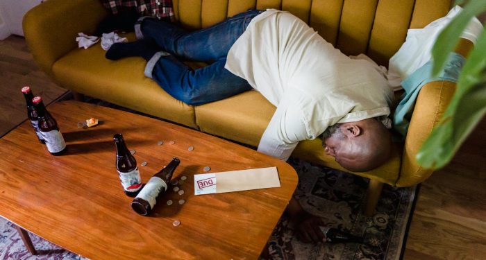 A man sleeping on a couch with empty bottles and an overdue bill, symbolizing financial stress and exhaustion.
