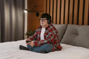 A young man with headphones and glasses playing video games on a bed, enjoying his leisure time.