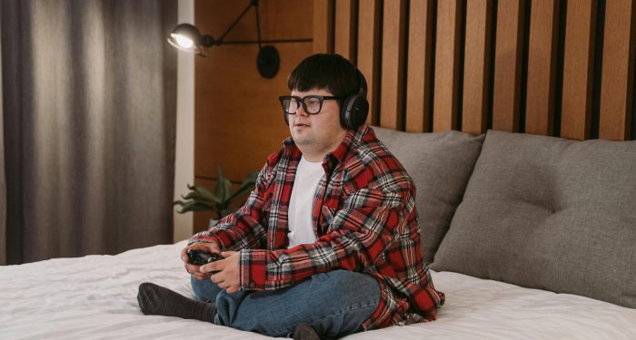 A young man with headphones and glasses playing video games on a bed, enjoying his leisure time.