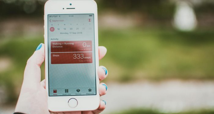 A person holds a smartphone showing a fitness app with step count and distance tracking outdoors.