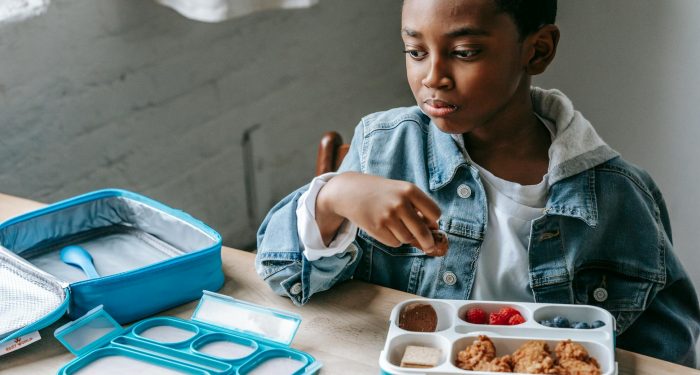 Crop contemplative African American schoolchild looking away at table with lunch container full of yummy food