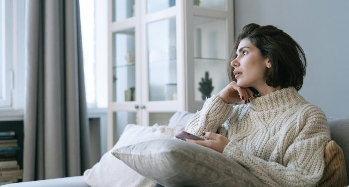 Woman in a comfy sweater sitting on couch, looking thoughtful with a smartphone in hand.