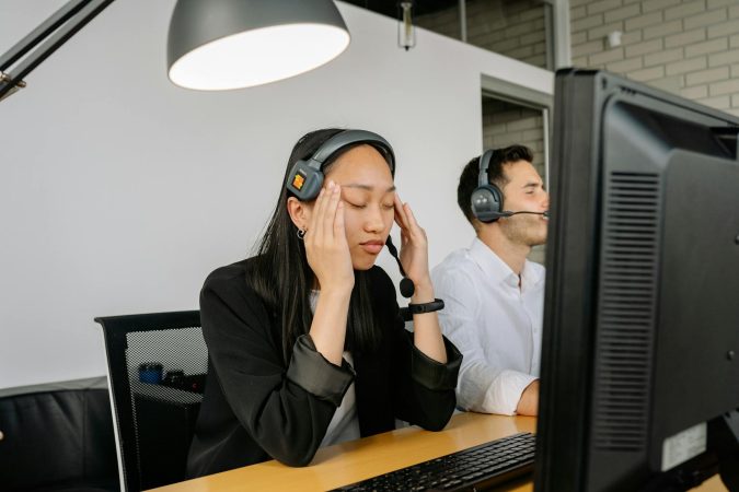 Asian woman at call center desk looking stressed, eyes closed, in office setting.