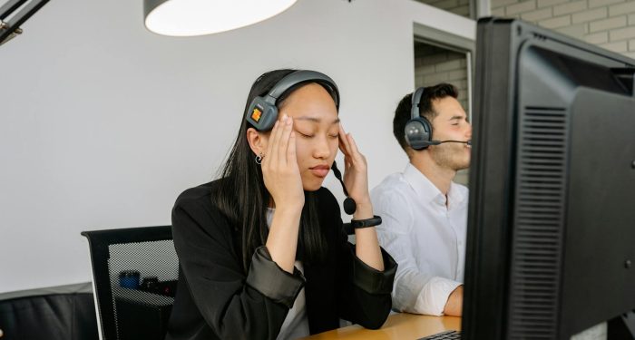 Asian woman at call center desk looking stressed, eyes closed, in office setting.