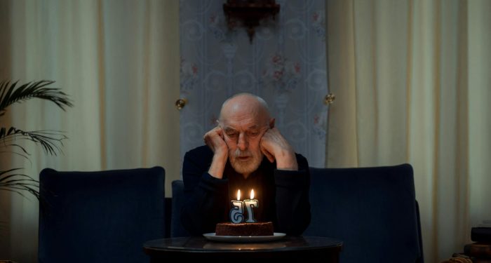 A senior man sitting thoughtfully by a lit birthday cake with a '75' candle indoors.