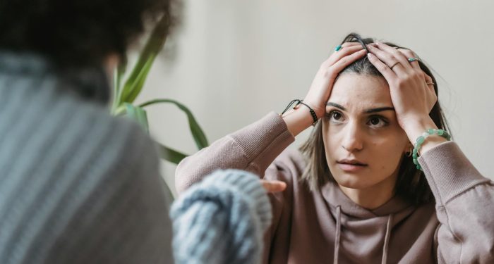 Worried female sitting near wall with hands on head and looking at faceless female while having conflict in light room
