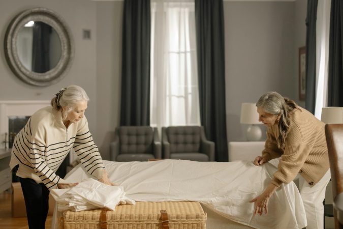 Two senior women folding sheets in a living room, preparing for moving out.