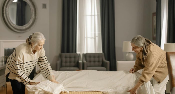 Two senior women folding sheets in a living room, preparing for moving out.