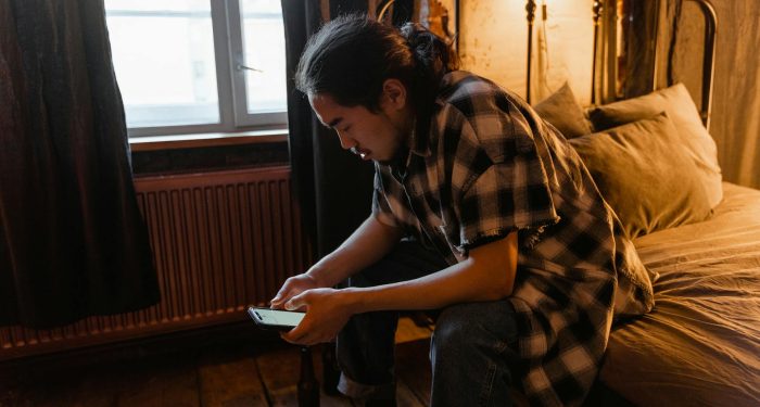 A young man with a contemplative expression sits on a bed looking at his phone inside a cozy room.