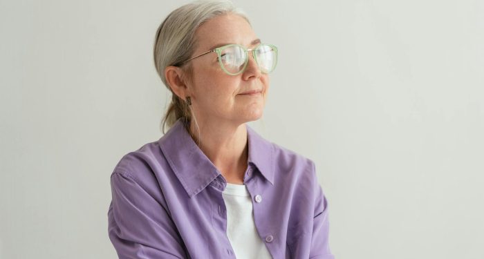 Senior woman with grey hair wearing eyeglasses and a purple shirt, looking thoughtful.