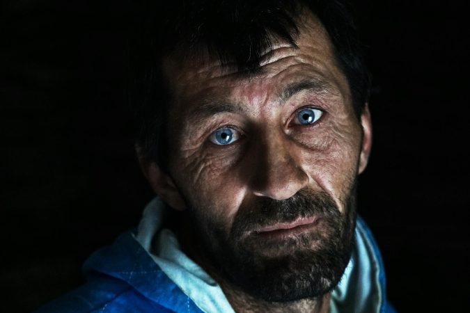 Close-up portrait of a man with blue eyes, beard, and a contemplative expression in dim lighting.