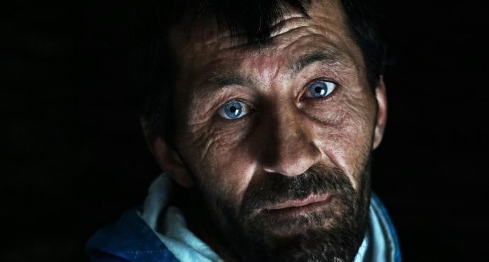 Close-up portrait of a man with blue eyes, beard, and a contemplative expression in dim lighting.