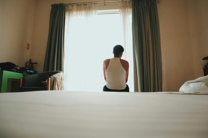 A man sits indoors facing a bright window, lost in thought, casting an introspective mood.